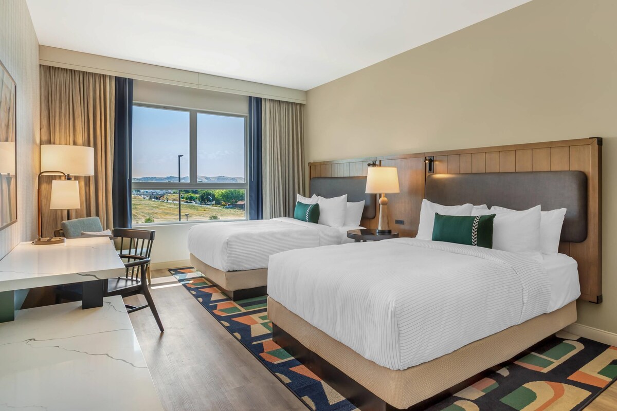 Quarto de hotel com duas camas, uma escrivaninha, uma cadeira e vista para o exterior.