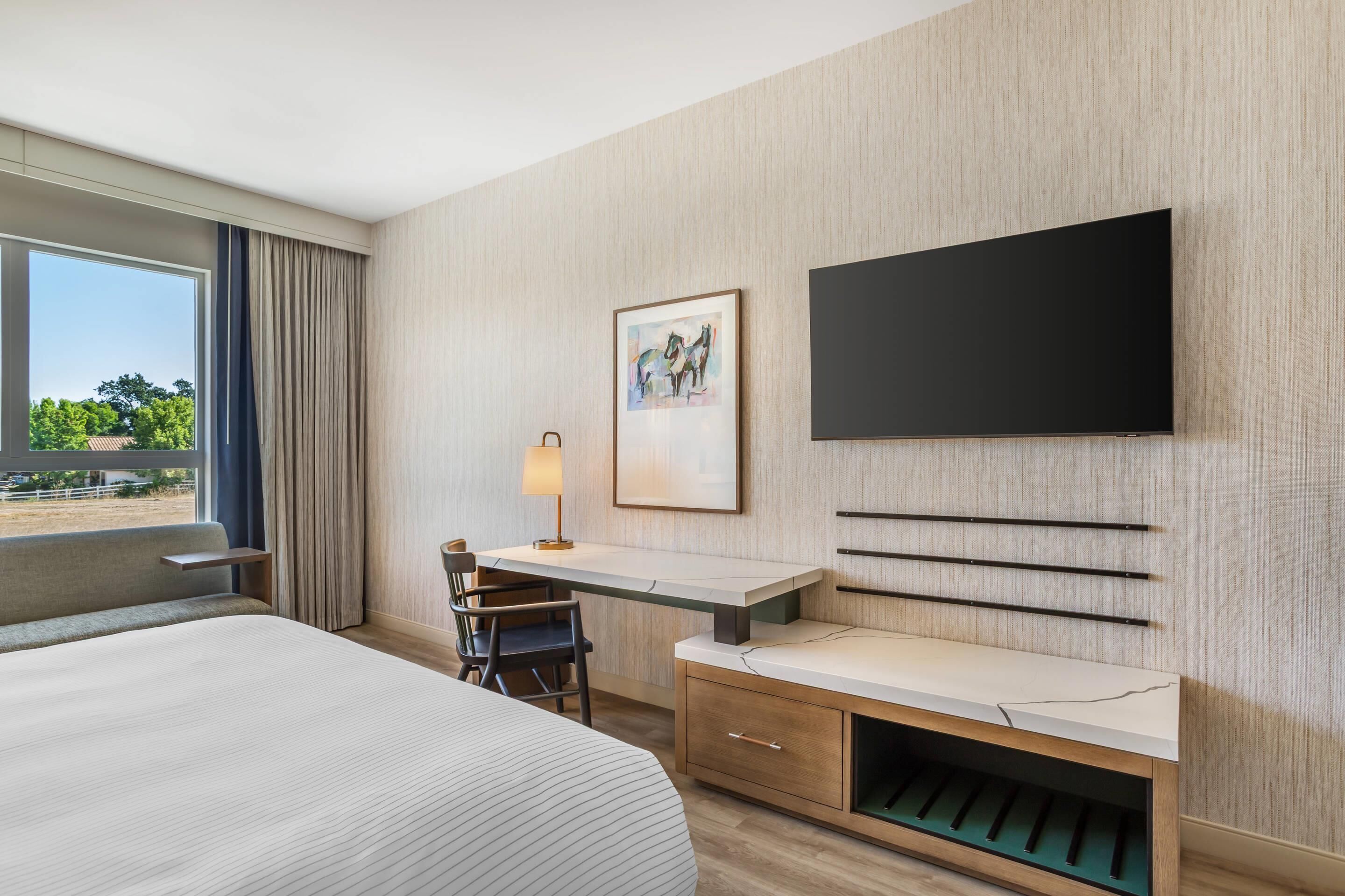 Hotelkamer met een groot bed, een bureau met stoel, een aan de muur bevestigde televisie en een raam met gordijnen.