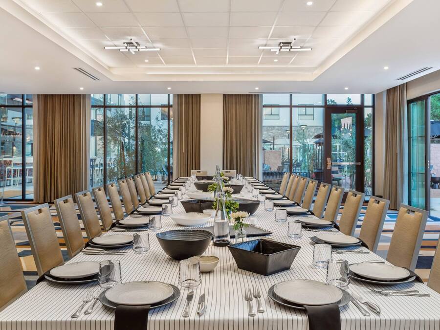 A long dining table set for a formal meal, with chairs on both sides, large windows, and a view of the outdoors.