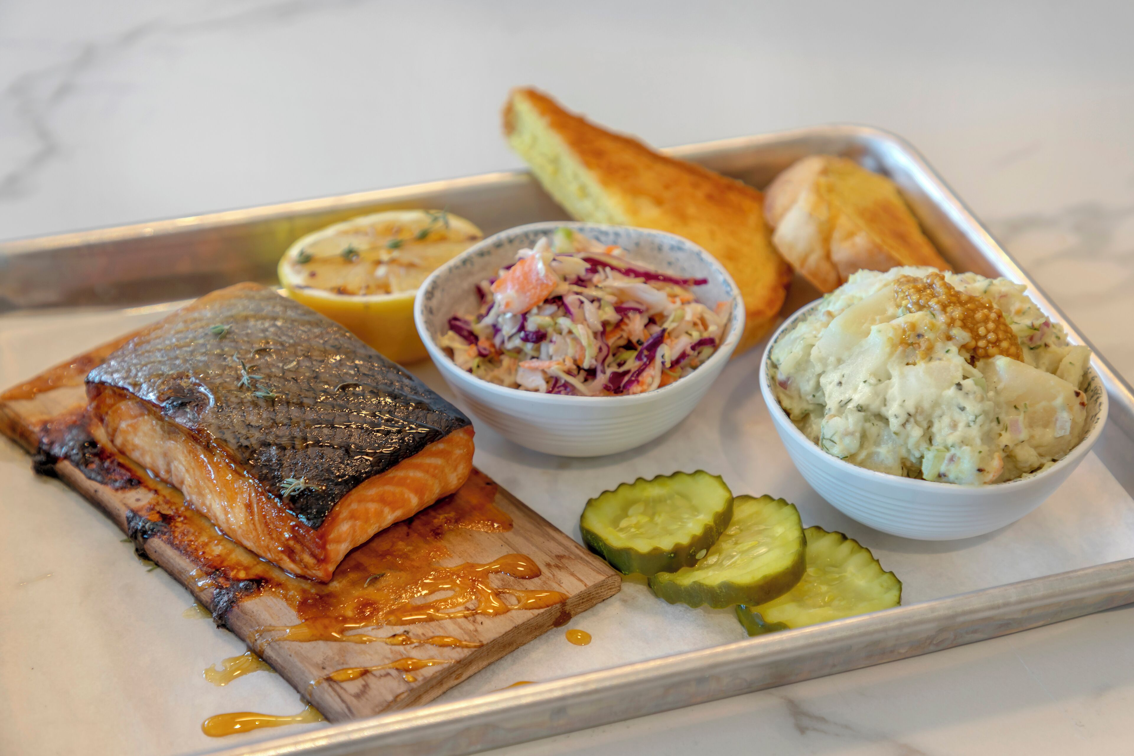 A tray with grilled salmon, coleslaw, potato salad, pickles, and a piece of bread.