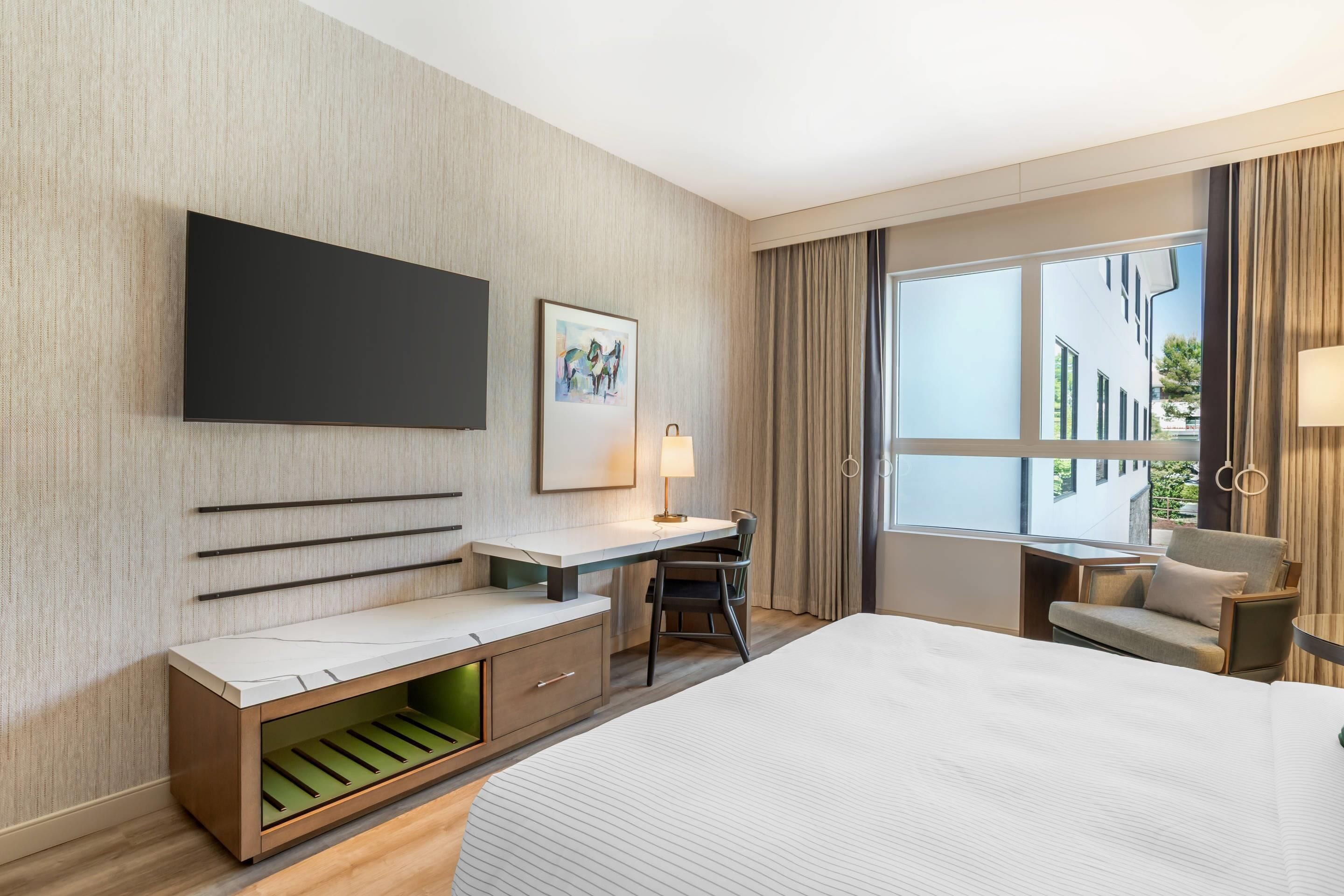 Een moderne hotelkamer met een groot bed, een bureau met stoel, een aan de muur bevestigde televisie en een groot raam met gordijnen.
