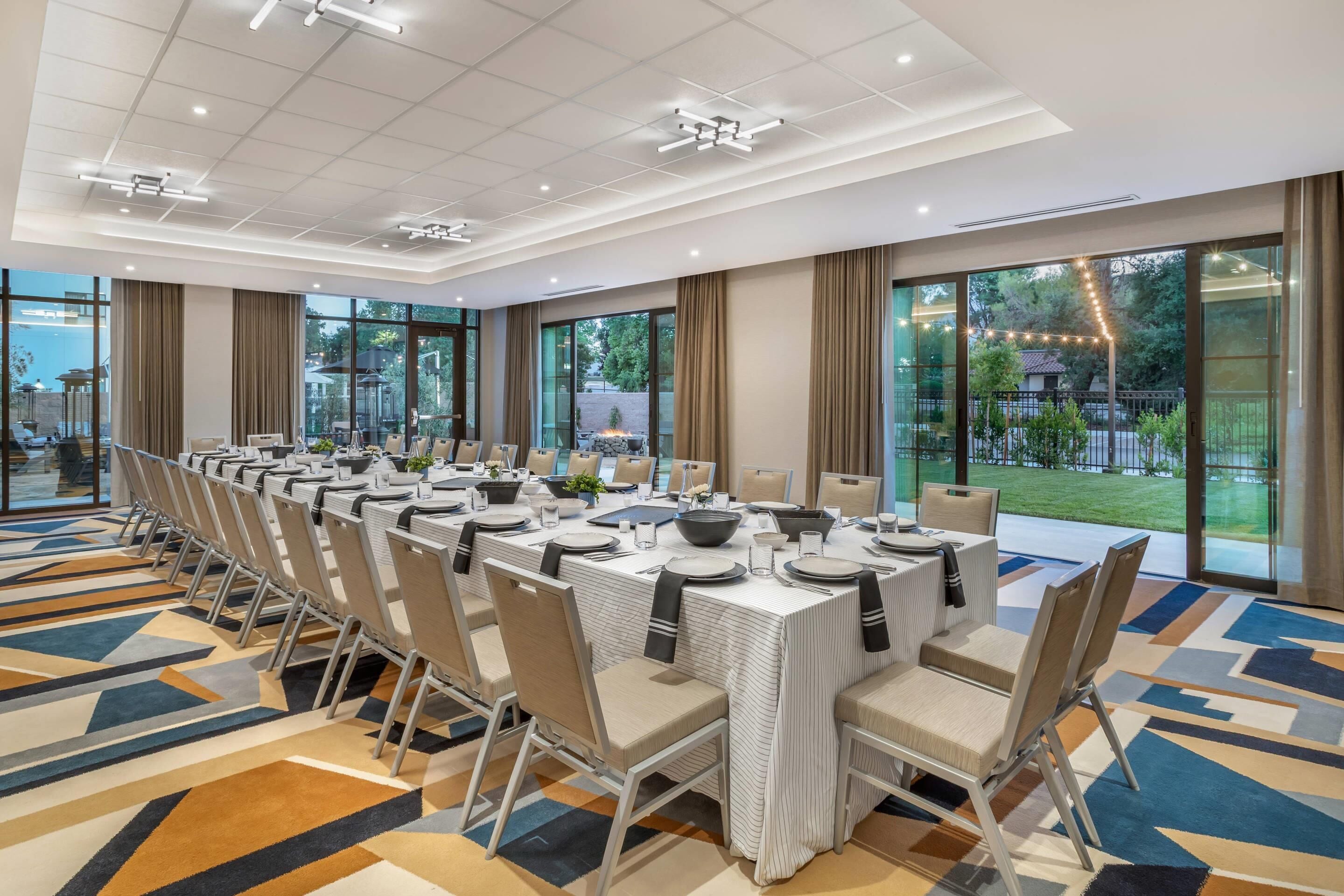 Un salón de conferencias con una larga mesa dispuesta para una reunión, ventanas amplias y vistas al exterior.