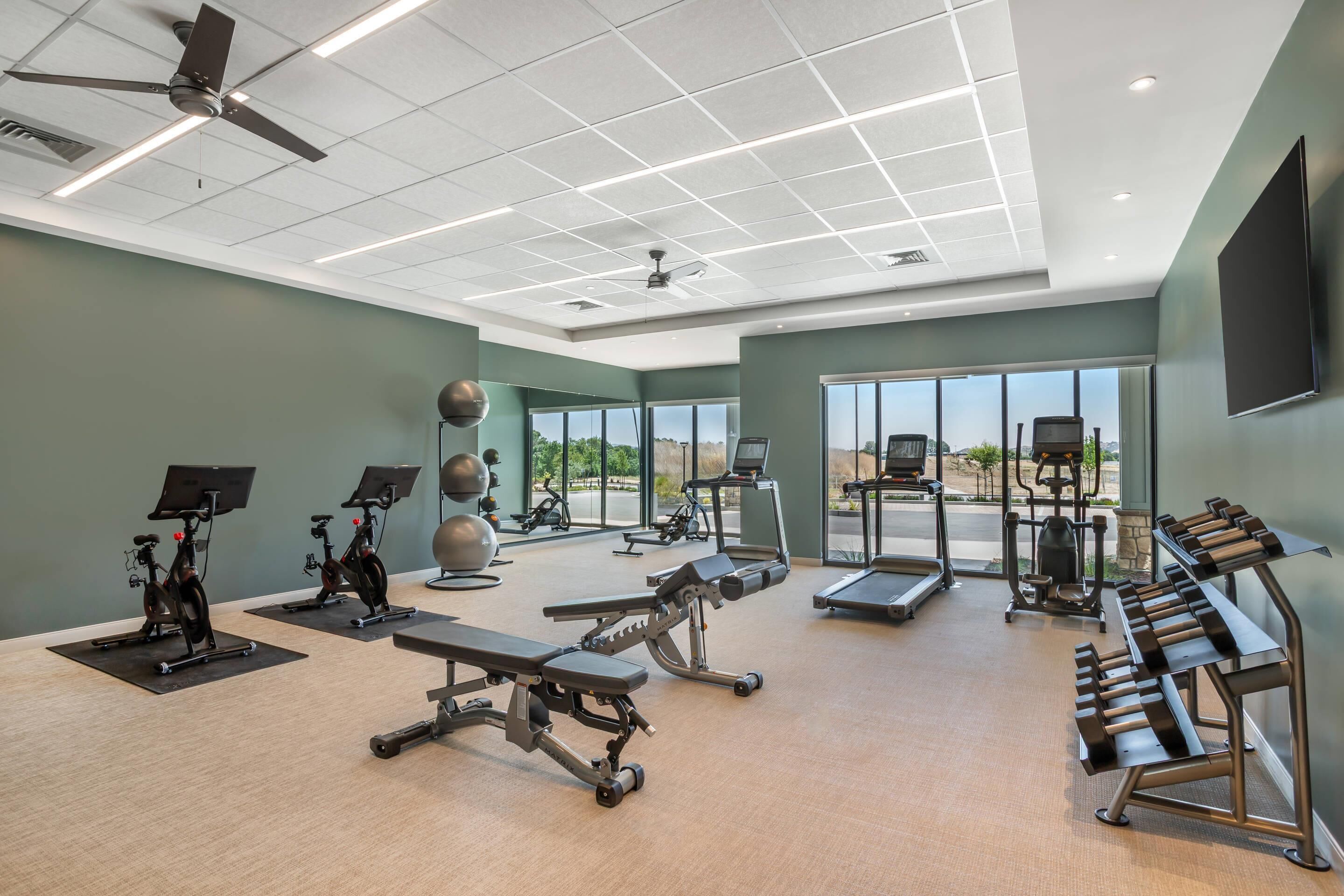 Une salle de sport moderne dotée de ventilateurs de plafond, d’appareils d’exercice et de grandes fenêtres offrant une vue sur l’extérieur.