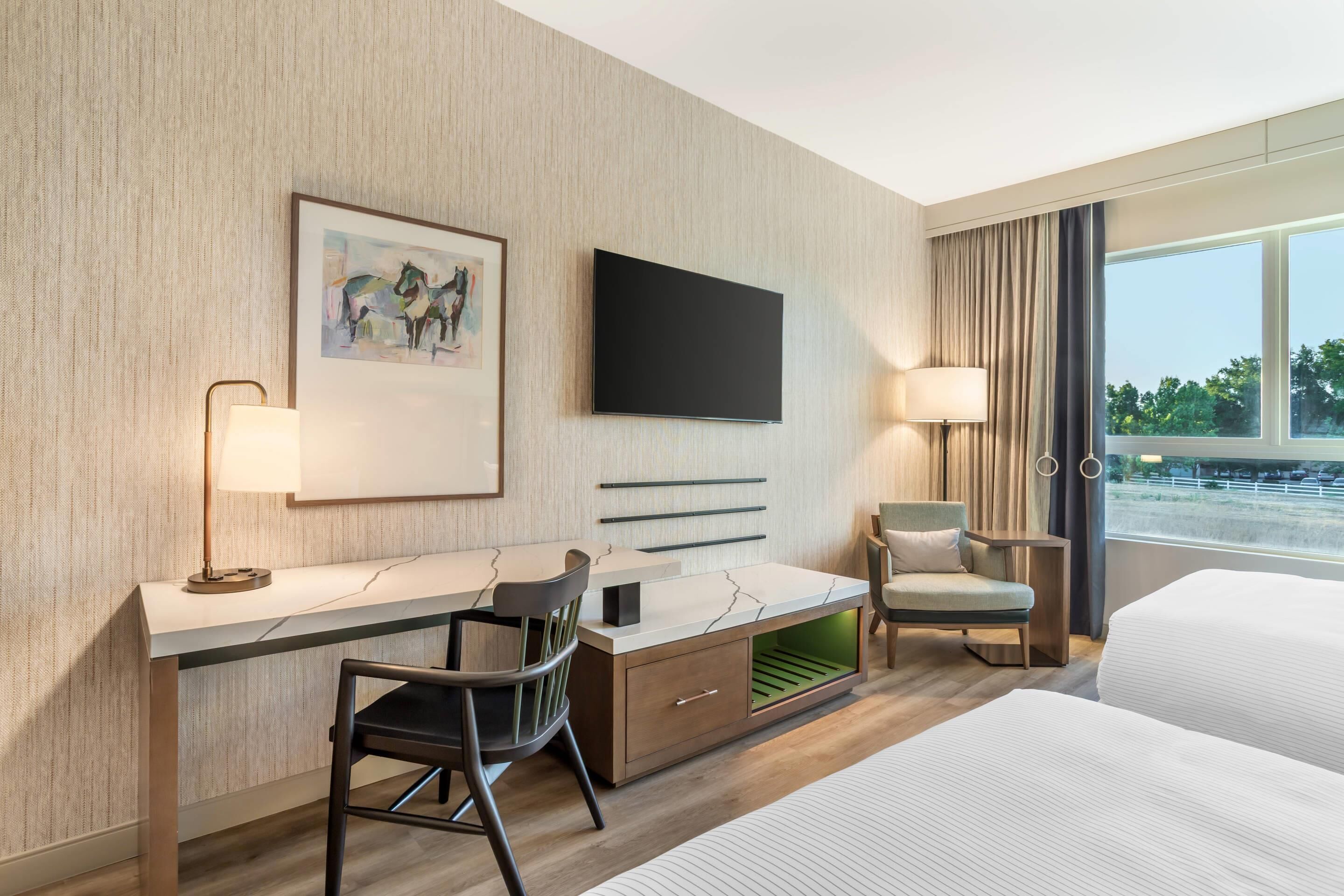 A modern hotel room with a large bed, a desk with a chair, a television, and a window with a view of the outdoors.