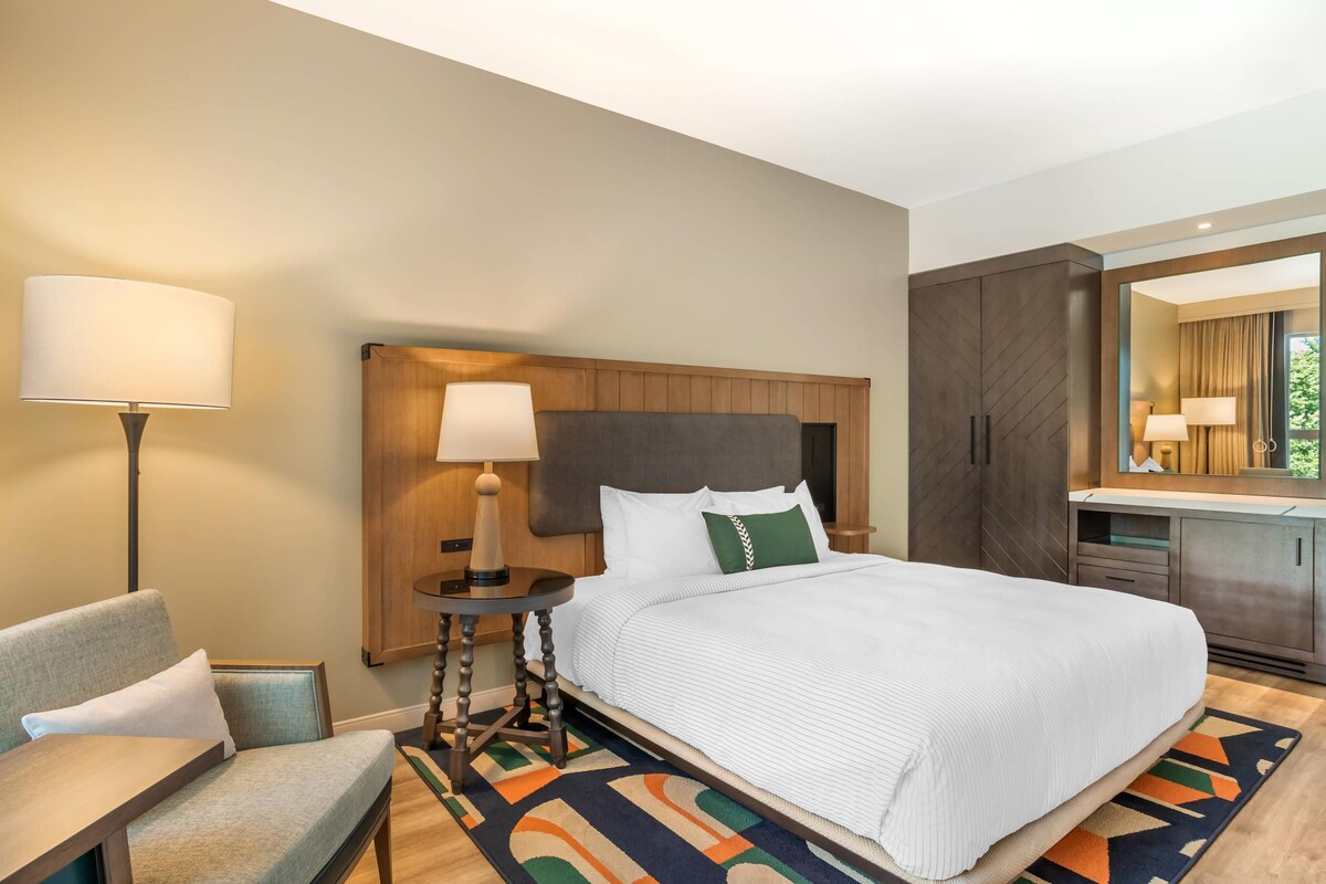 A hotel room with a bed, a desk, a chair, a lamp, a dresser, and a mirror.