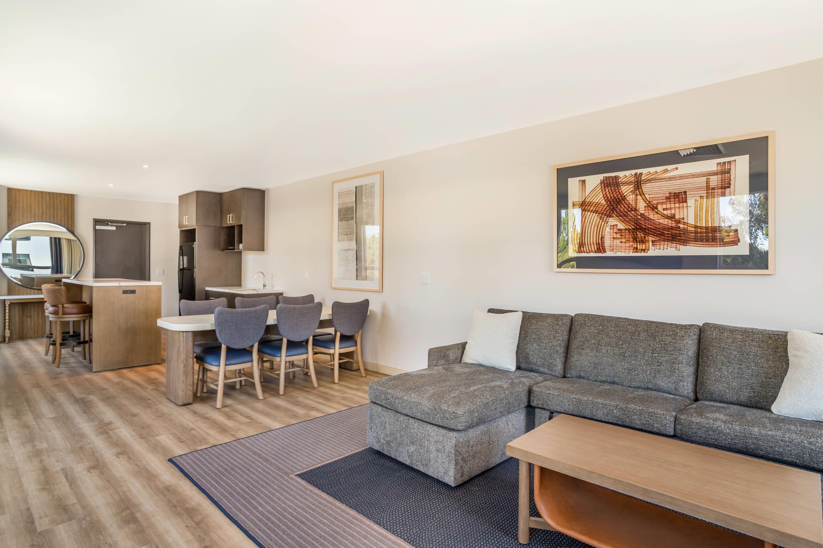 A modern living room with a grey sectional sofa, a dining area with chairs, a kitchen area, and a large abstract painting on the wall.