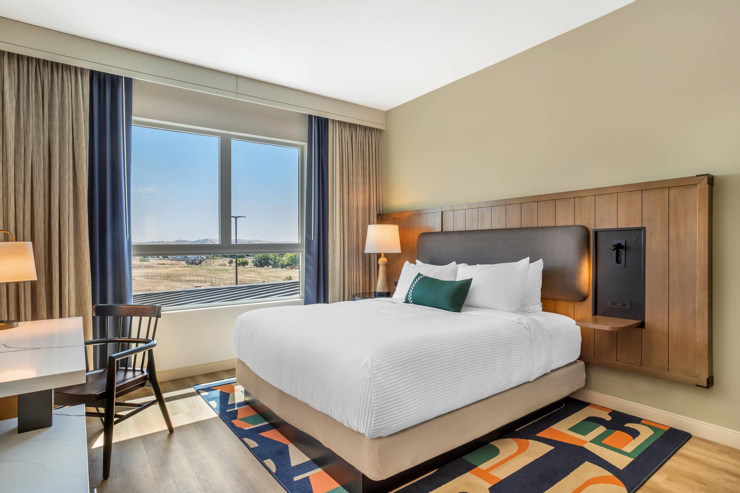 A hotel room with a large bed, a desk, a chair, a lamp, and a view of the outdoors.