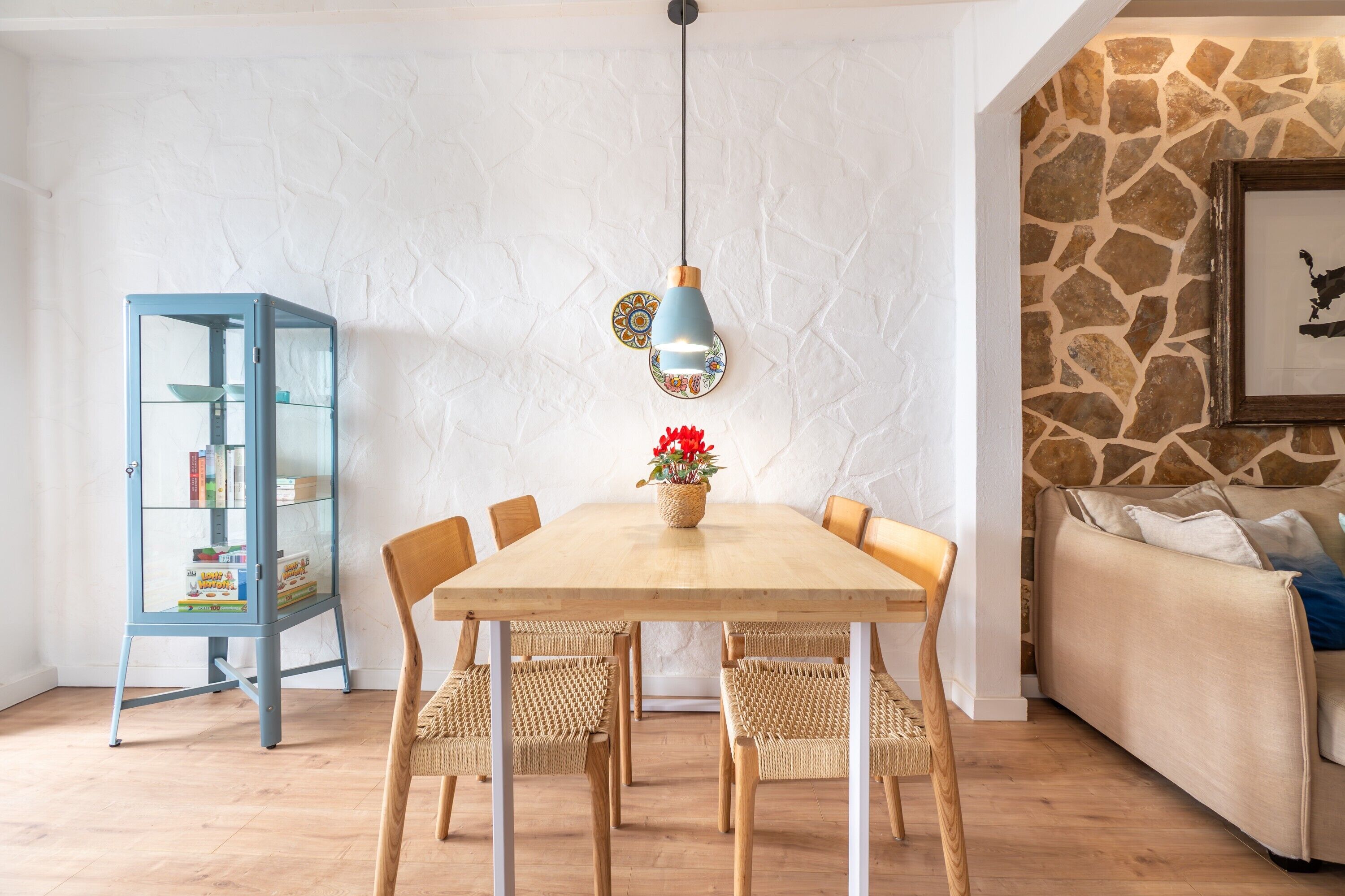 Un coin repas avec une table en bois, des chaises en osier, un buffet bleu aux portes vitrées et un mur en pierre.