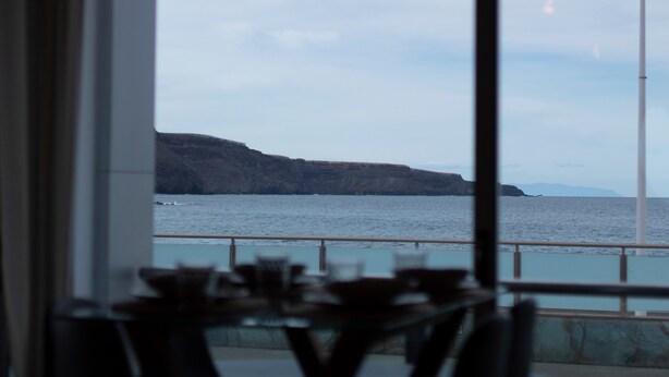 A view through a window of a coastal area with a railing, a table, and chairs in the foreground.