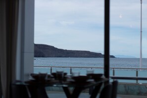 A view through a window of a coastal area with a railing, a table, and chairs in the foreground.