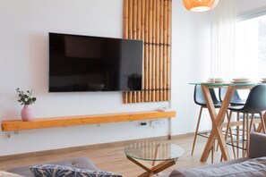 A modern living room with a flat-screen TV, bamboo wall decor, a glass coffee table, and a dining area with chairs and a table.