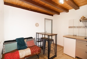 Una habitación con techo de madera, una cama con manta estampada, una mesita, una silla y una pequeña cocina.