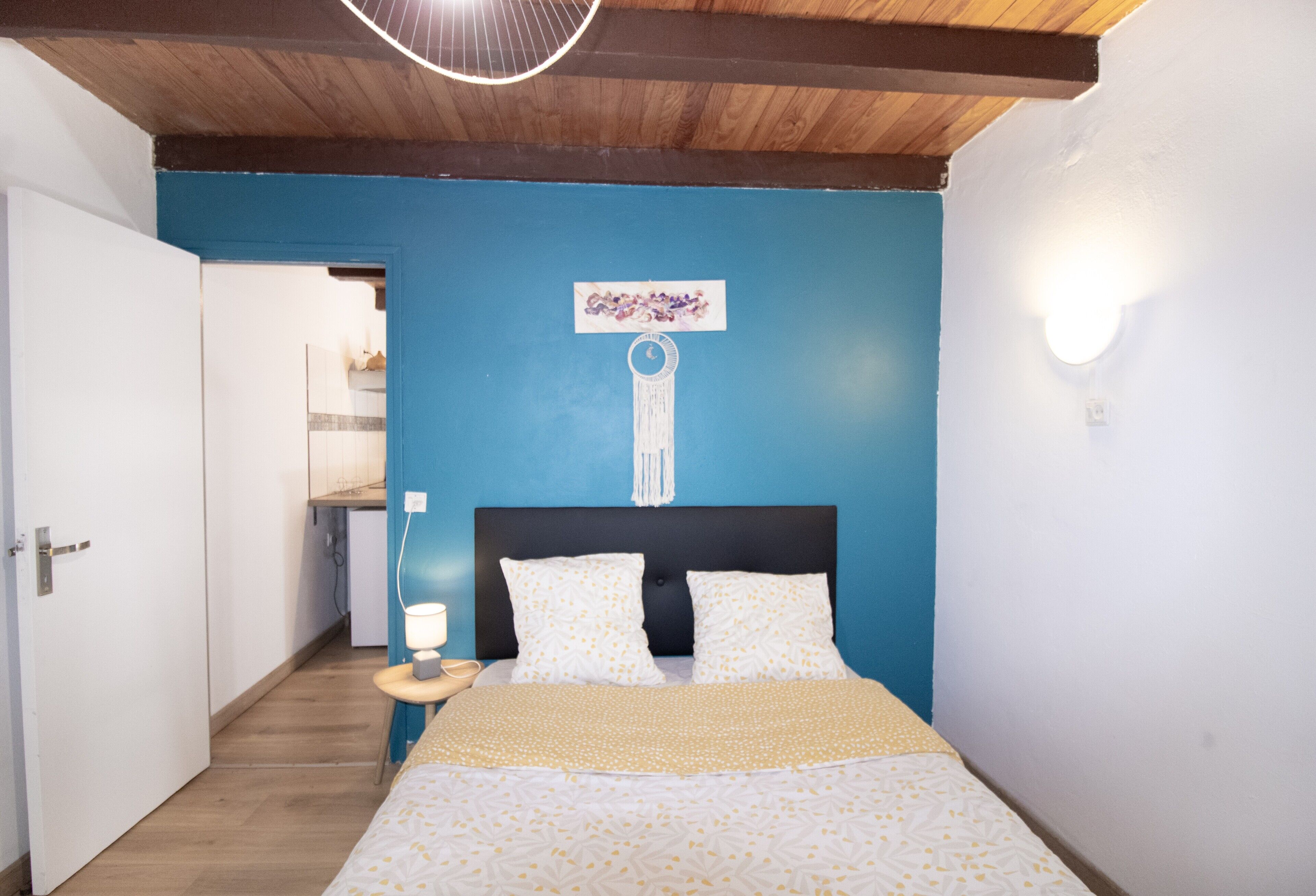 A bedroom with a bed, a nightstand, a wall-mounted light, and a door leading to another room.