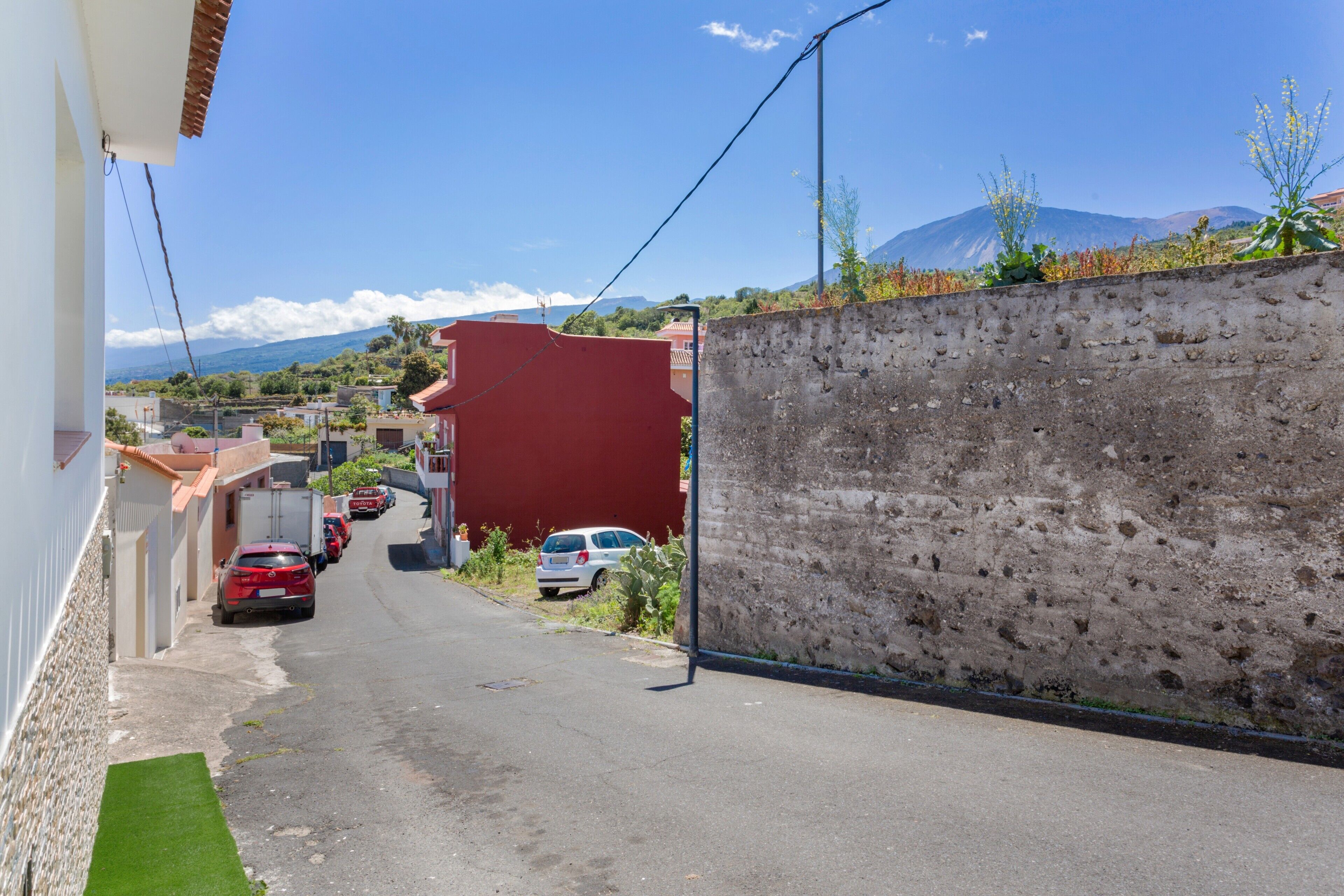 Une rue étroite bordée de voitures garées, un immeuble rouge et un mur en béton.