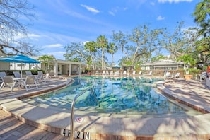 A swimming pool with lounge chairs and umbrellas, surrounded by trees and a covered area.