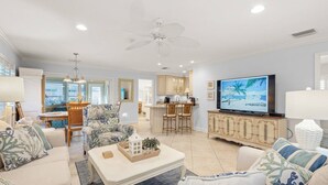 A spacious living room with a large TV, comfortable seating, a kitchen area, and a ceiling fan.