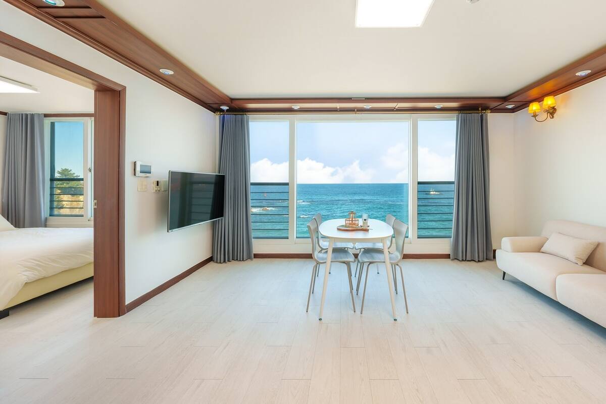 Une chambre d’hôtel moderne dotée d’une grande fenêtre donnant sur la mer, d’un téléviseur à écran plat fixé au mur, d’une table à manger ronde avec des chaises et d’un canapé beige.