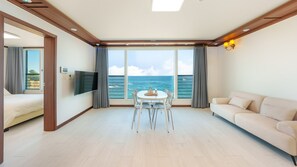 Una habitación de hotel moderna con un ventanal que ofrece vista al mar, un televisor de pantalla plana en la pared, una mesa de comedor redonda con sillas y un sofá color beige.