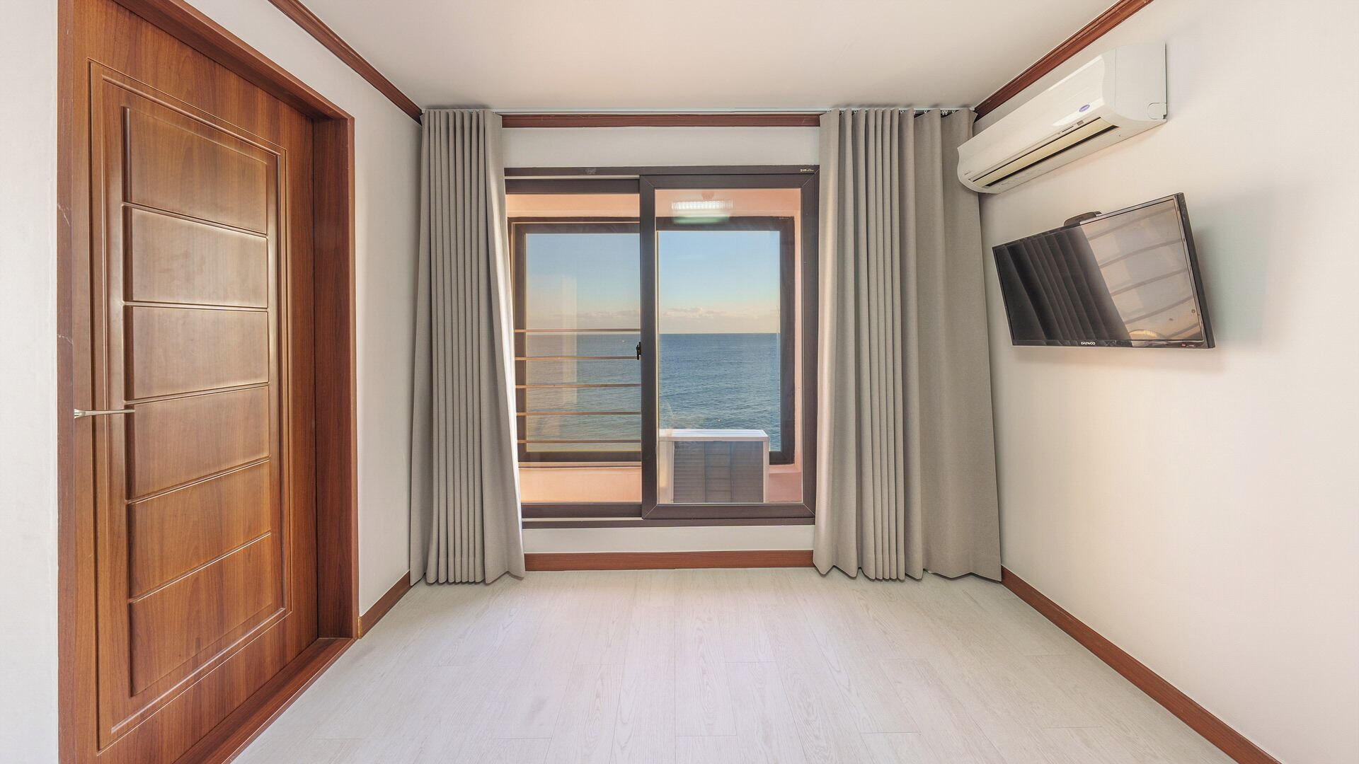 Een hotelkamer met een houten deur, een schuifglasdeur naar een balkon, een airconditioner en een televisie aan de muur.
