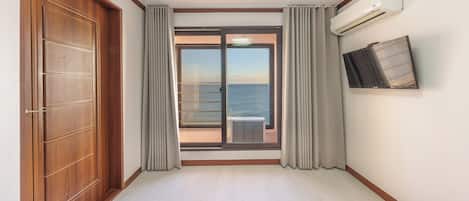Una habitación de hotel con puerta de madera, puerta corredera de vidrio a un balcón, unidad de aire acondicionado y televisor montado.