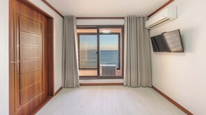 Une chambre d’hôtel avec une porte en bois, une porte-fenêtre coulissante donnant sur un balcon, un appareil de climatisation et un téléviseur fixé au mur.