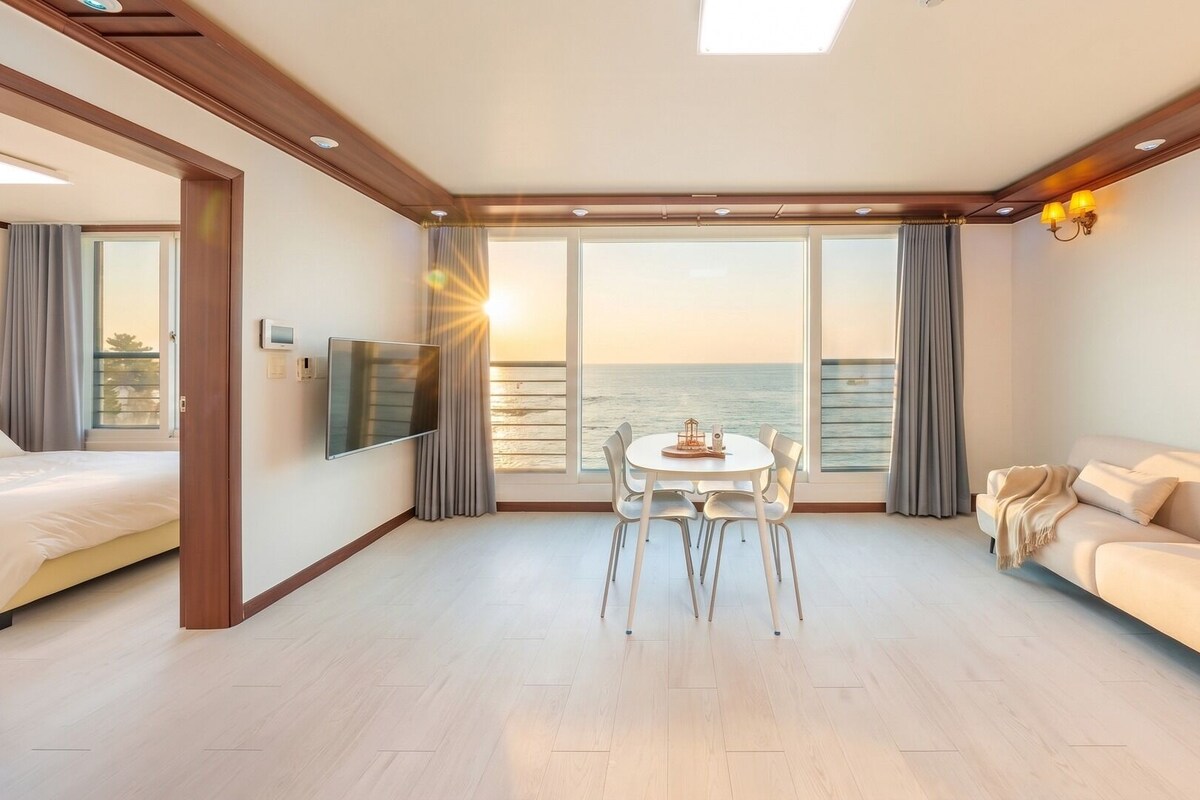 Une chambre d’hôtel moderne dotée d’une grande fenêtre donnant sur la mer, d’un coin repas avec une table et des chaises, d’un canapé et d’un téléviseur à écran plat fixé au mur.