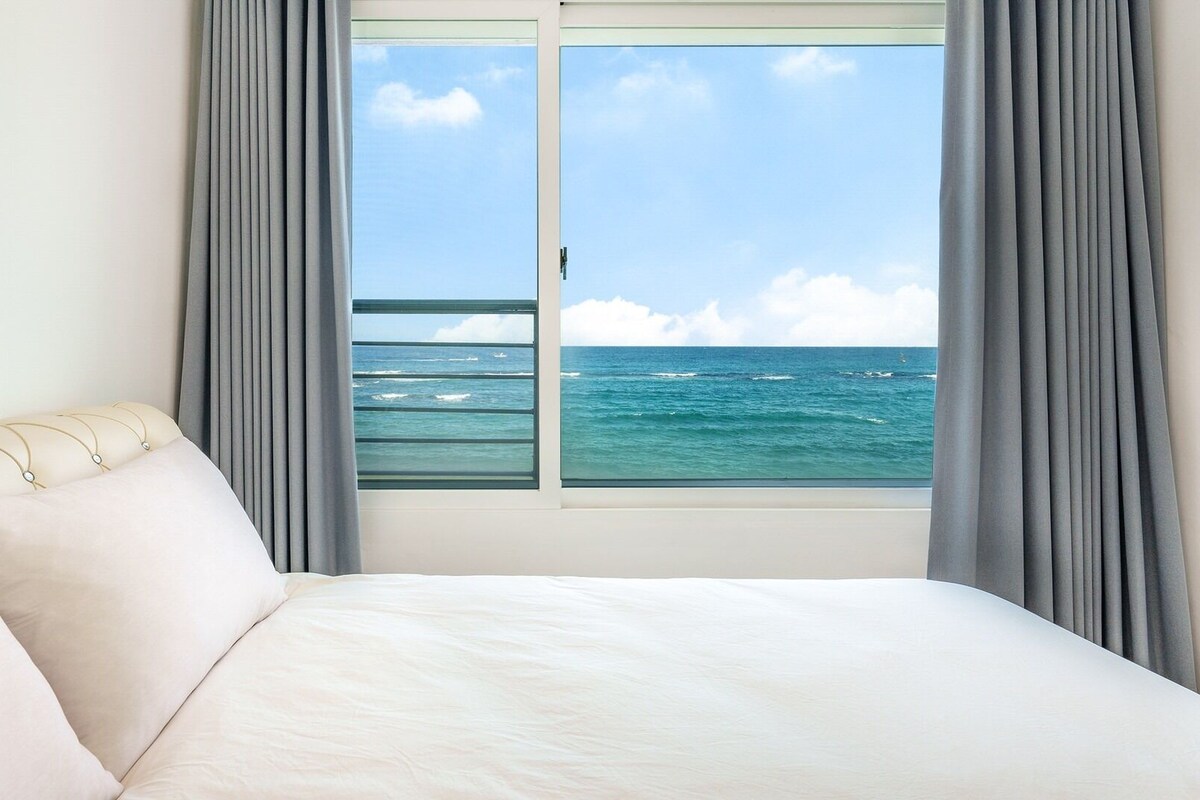 Une chambre d’hôtel avec un lit, une fenêtre donnant sur l’océan et des rideaux gris.