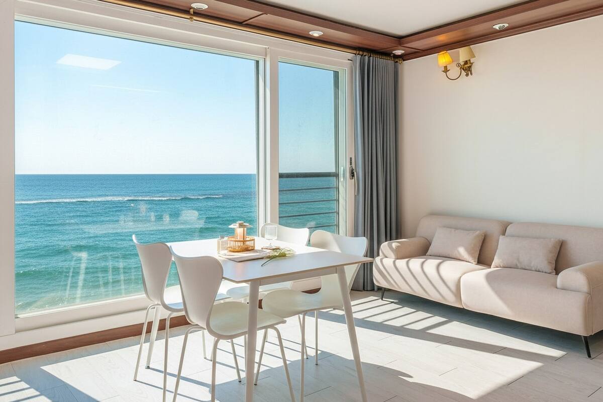 Une chambre moderne avec une grande fenêtre donnant sur la mer, une table blanche avec des chaises et un canapé beige.