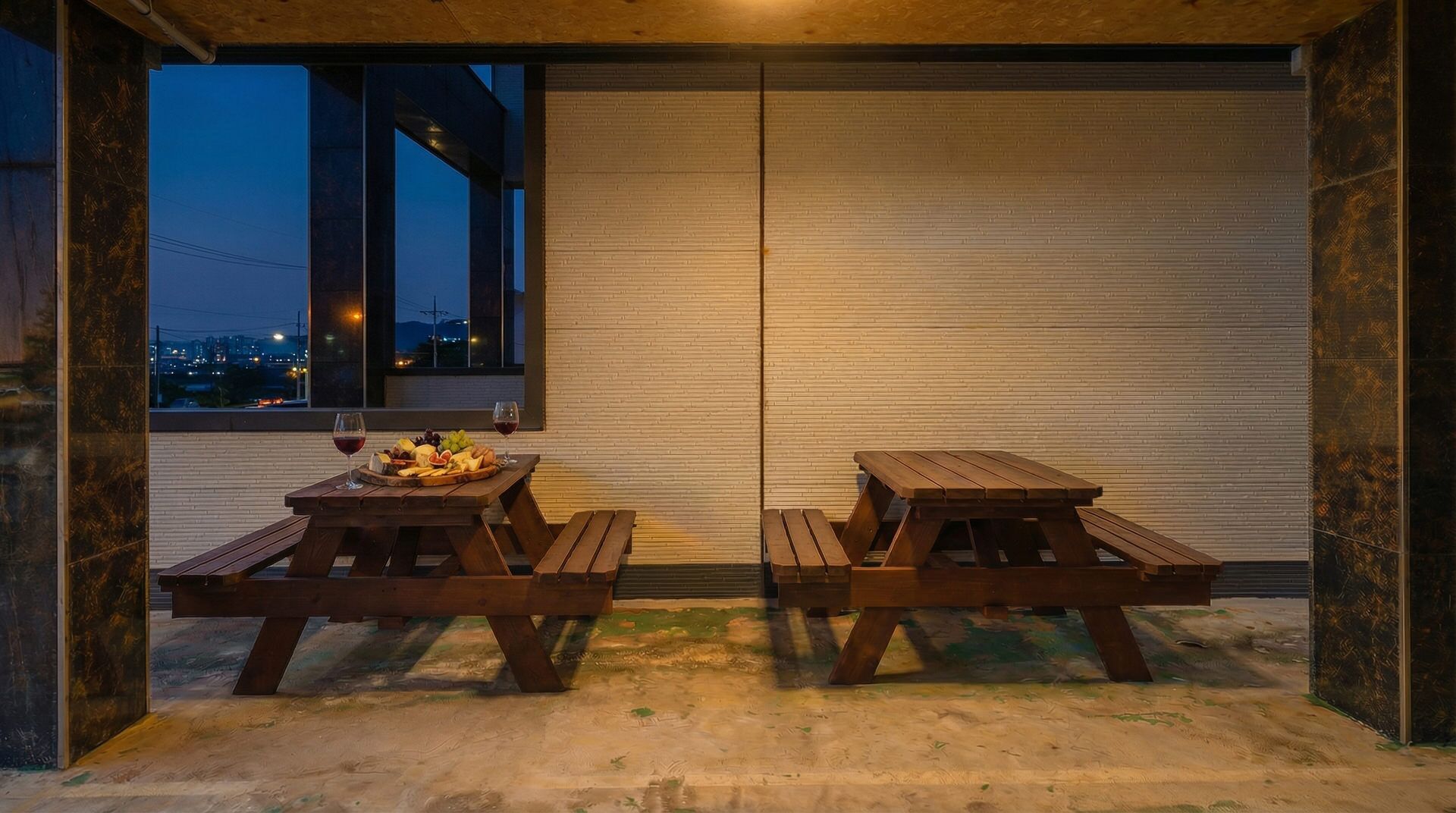 A patio with two wooden picnic tables, one with a fruit and cheese board and wine glasses, and a cityscape view at dusk.