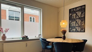 A modern dining area with a round wooden table, blue chairs, a vase with flowers, and a cityscape painting on the wall. - Cozy Flats Silkeborg - S2 (Silkeborg)