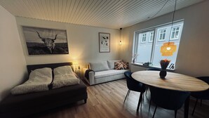 A modern living room with a sofa, a dining table with chairs, a lamp, and a window with a view of buildings. - Cozy Flats Silkeborg - S2 (Silkeborg)