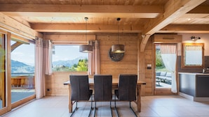 Ambiente interno in legno con tavolo da pranzo, sedie, angolo cottura e vista sulle montagne.