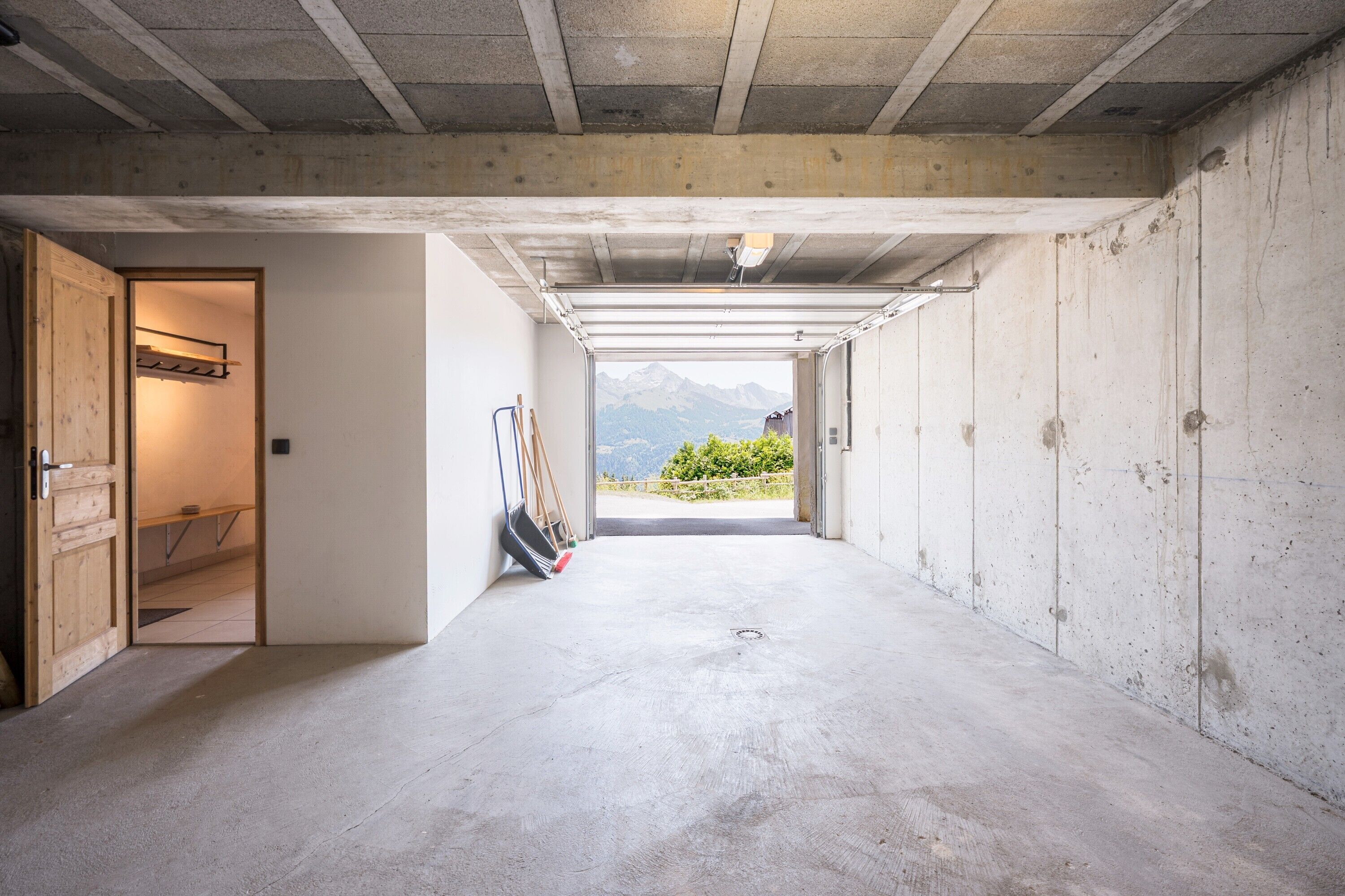 Ein Zimmer mit Betonboden, Holztür, einem an der Wand angelehnten Besen und Blick auf Berge durch eine offene Tür.