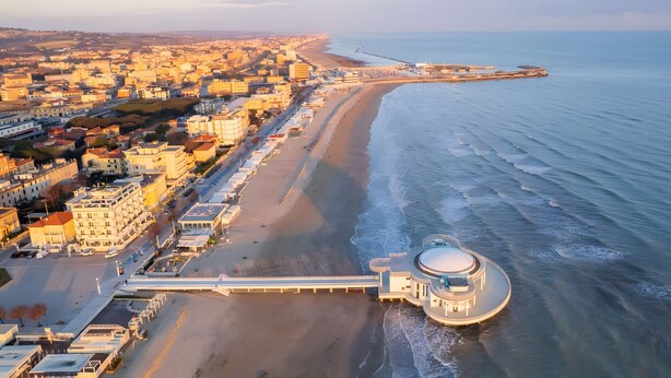 Une vue aérienne d’une ville côtière, avec un imposant bâtiment circulaire situé près de la plage, une longue jetée s’étendant dans l’eau et une zone urbaine dense comprenant divers bâtiments.