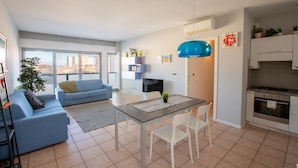 Un appartement moderne comprenant un salon, un coin repas et une cuisine. On y trouve une grande fenêtre, un canapé bleu, une table à manger avec des chaises, ainsi qu’une cuisine équipée de placards et d’une cuisinière.