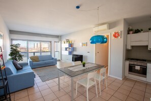 Un apartamento moderno con sala de estar, comedor y cocina. Cuenta con un ventanal grande, un sofá azul, una mesa de comedor con sillas y una cocina con armarios y estufa.