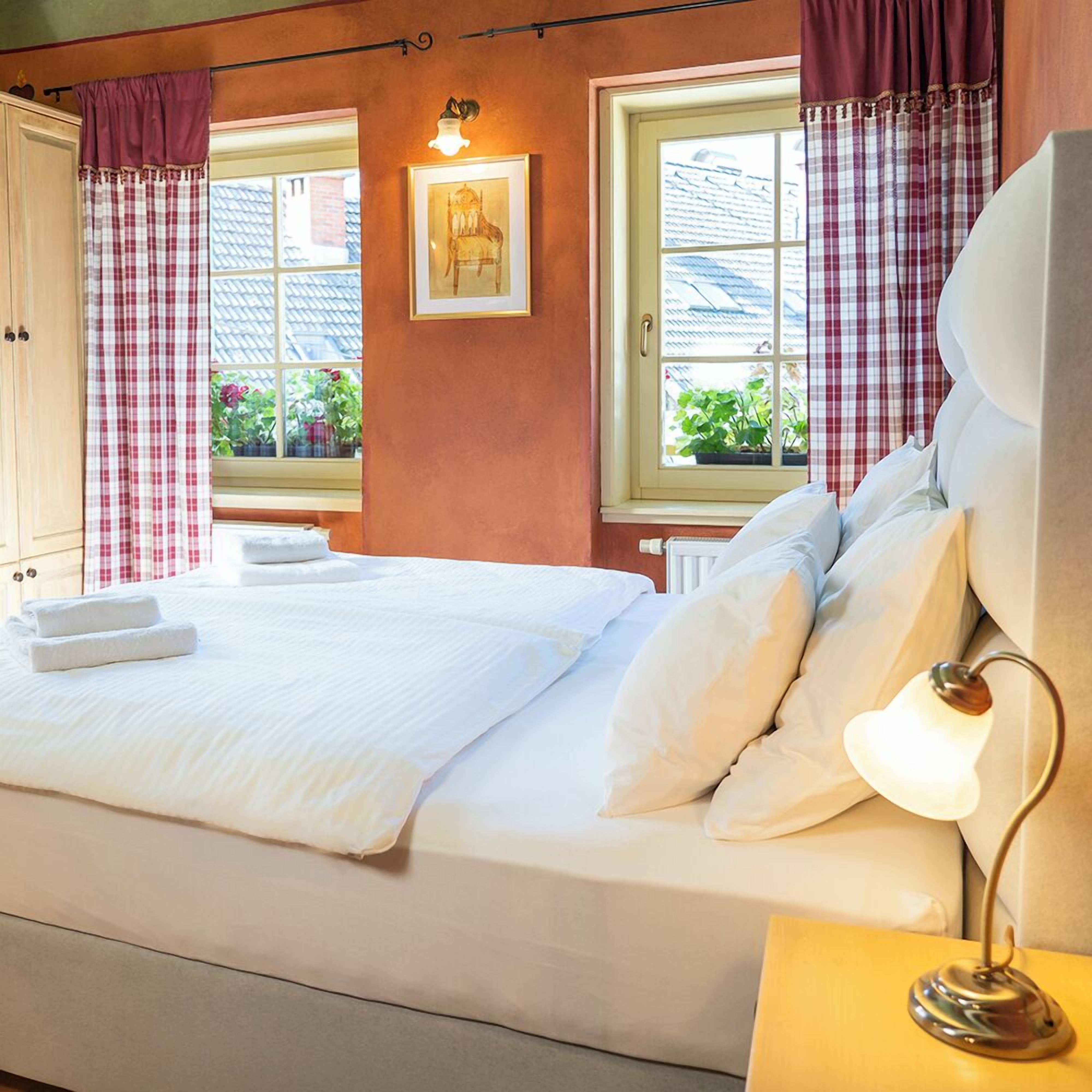 A neatly made bed with white linens, a bedside lamp, and a window with checkered curtains.