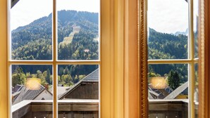 Uitzicht door een raam met houten kader, waarop een bergachtig landschap met bomen en gebouwen te zien is.