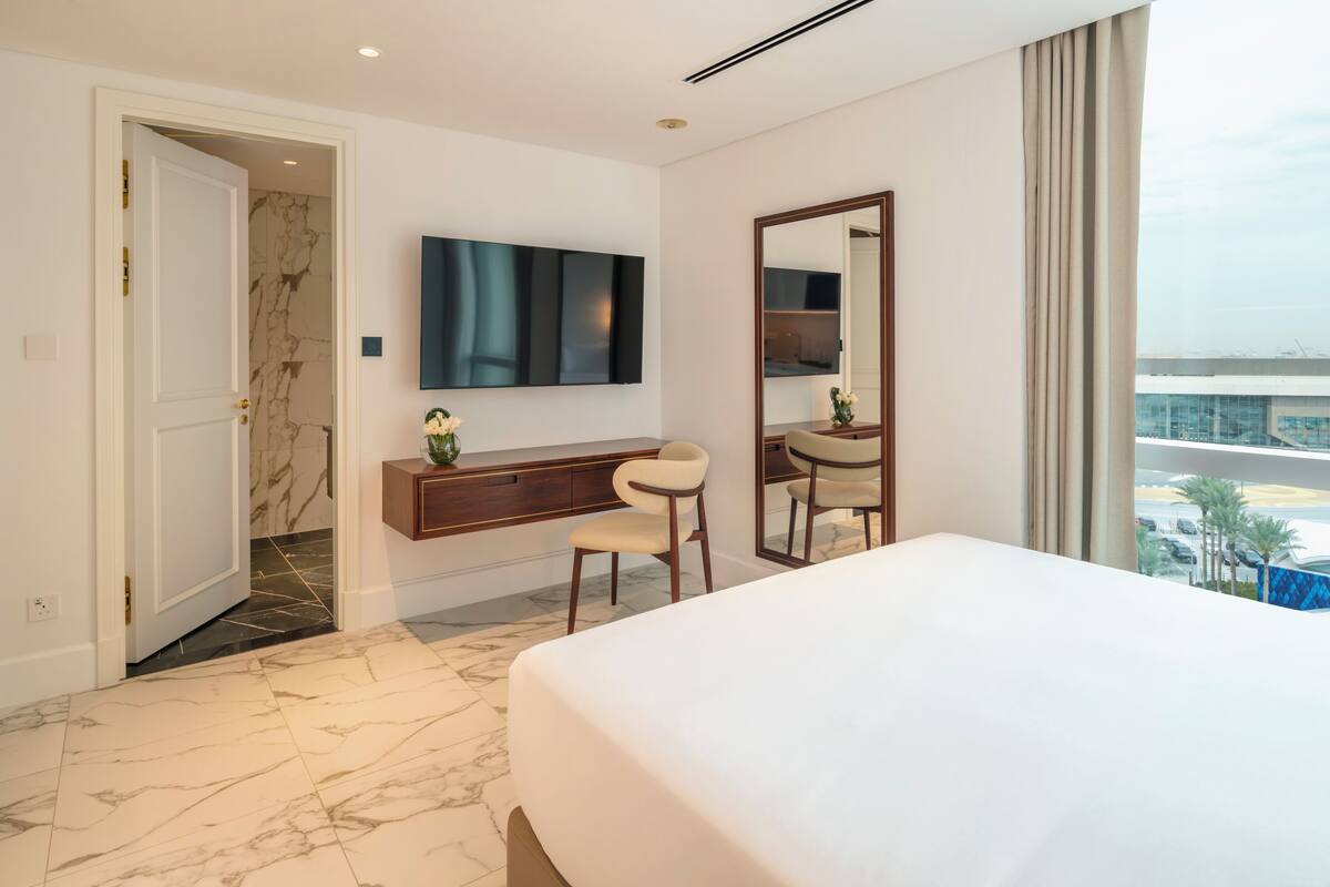 A hotel room with a large bed, a TV mounted on the wall, a vanity with a mirror, and a view of the cityscape through the window.