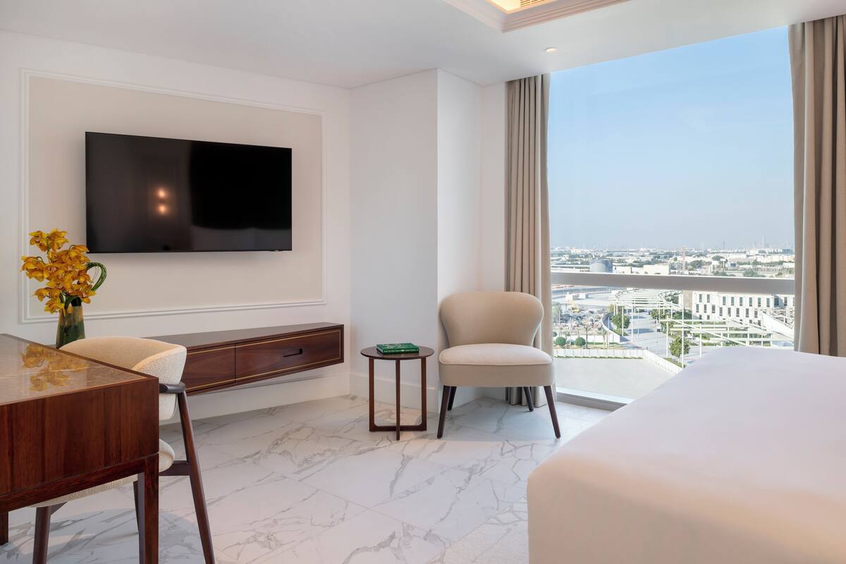 A hotel room with a large bed, a TV mounted on the wall, a small table with a vase of flowers, a chair, and a view of the city through large windows.