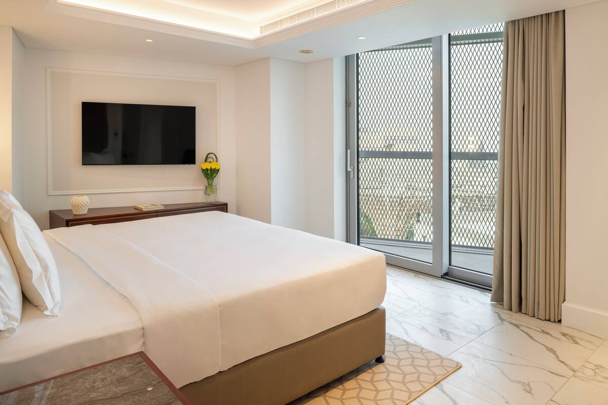 A modern hotel room with a large bed, a flat-screen TV mounted on the wall, a small table with a vase of flowers, and a sliding glass door leading to a balcony.