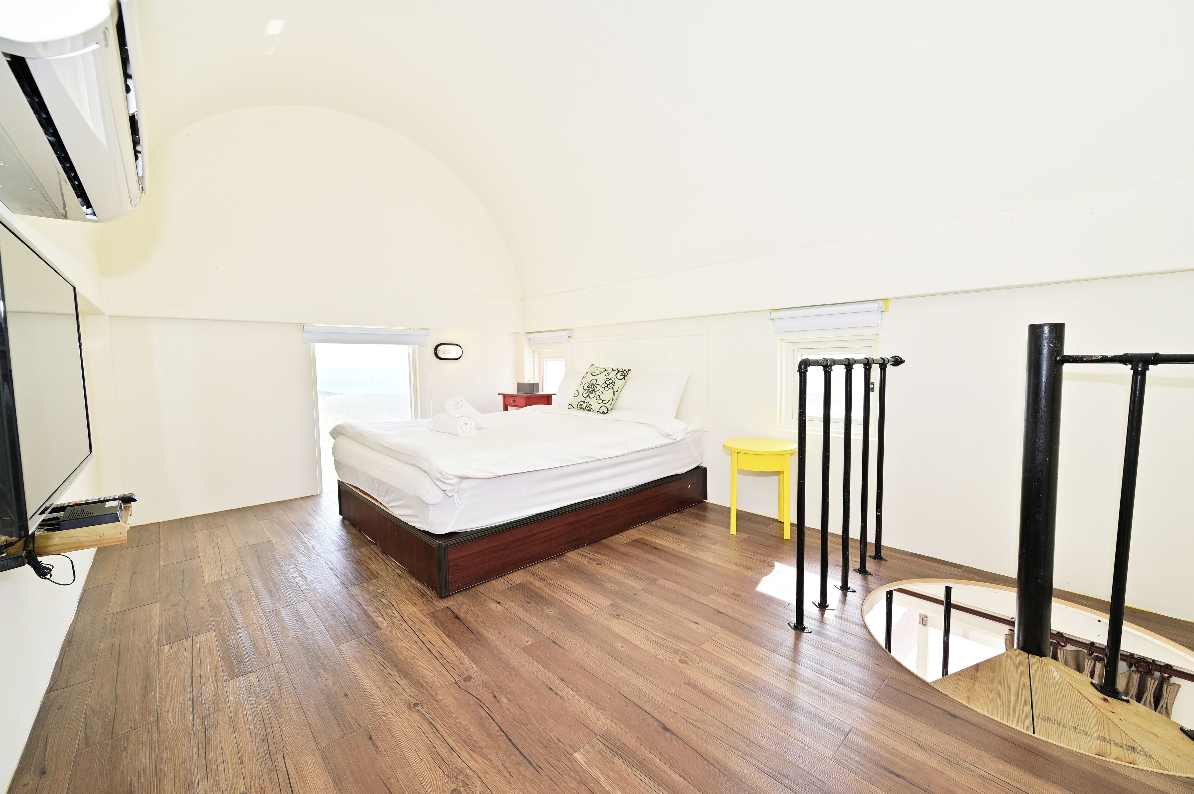 A bedroom with a wooden floor, a bed, a small yellow table, and a staircase with a railing.