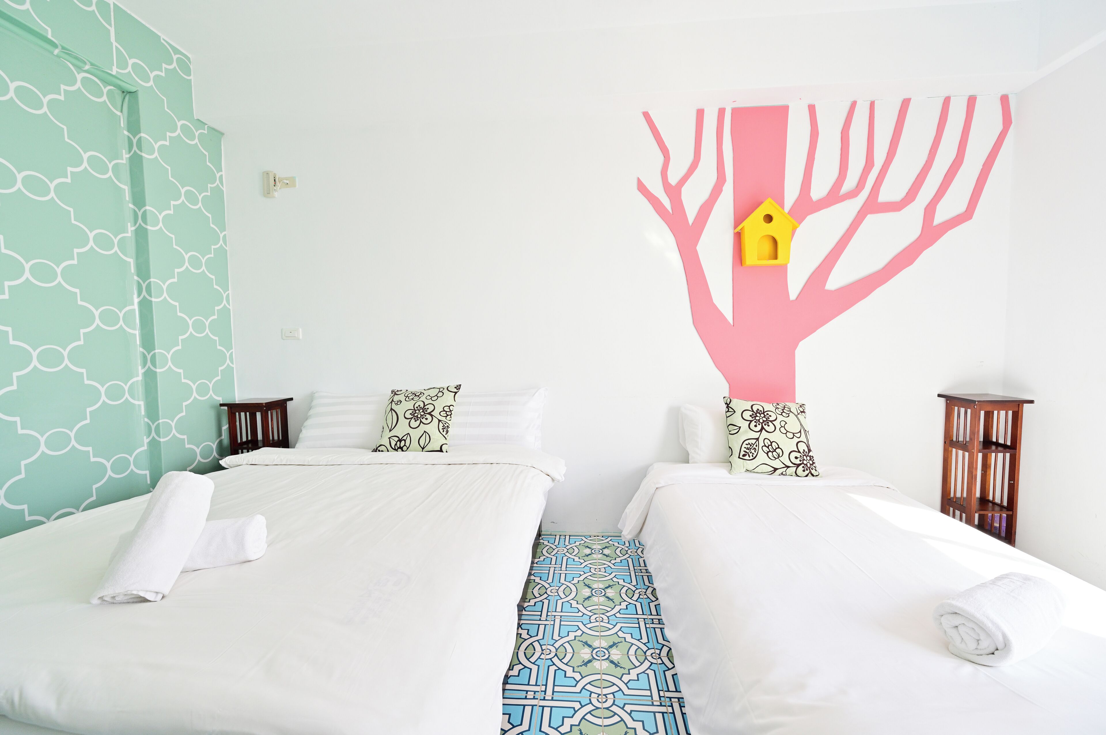 Une chambre d’hôtel avec deux lits, chacun recouvert d’une courtepointe blanche et agrémenté d’une serviette blanche roulée. On y trouve une table de chevet en bois avec une lampe d’un côté, et une décoration murale représentant un arbre avec une nichoir.