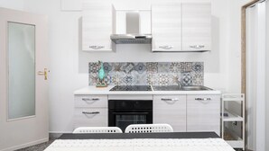 Una cocina compacta con armarios blancos, salpicadero de azulejos, horno integrado y un pequeño comedor con dos sillas.