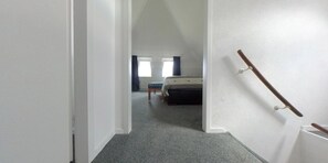 A narrow hallway leading to a bedroom with a bed, a window with curtains, and a wooden handrail on the right.