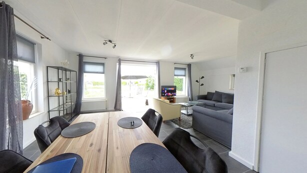 A modern living room with a wooden dining table, black chairs, a grey sofa, and a TV. - Topsleep Villa Lathum (Watervogel 12, 6988 CB LATHUM)