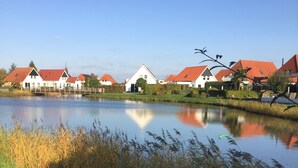 A serene lakeside view with traditional houses, a wooden bridge, and surrounding vegetation. - Topsleep Villa Lathum (Watervogel 12, 6988 CB LATHUM)