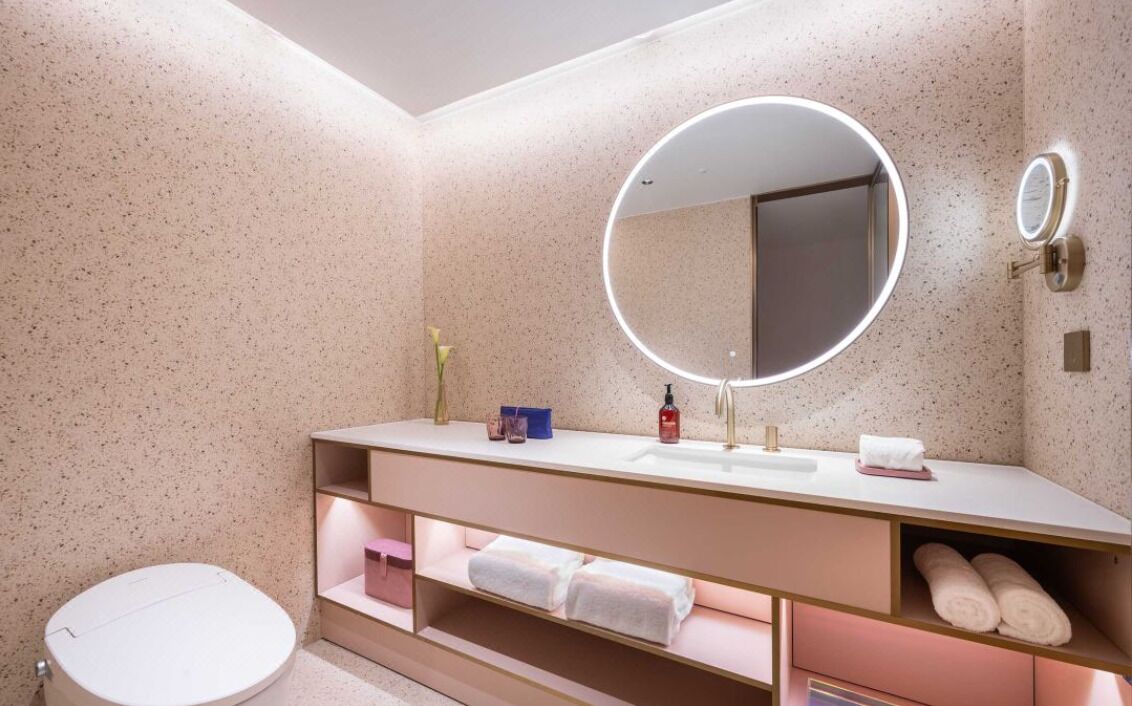 Un baño moderno con un espejo redondo, un lavabo blanco y un inodoro. Hay estantes con toallas y una pequeña caja azul.