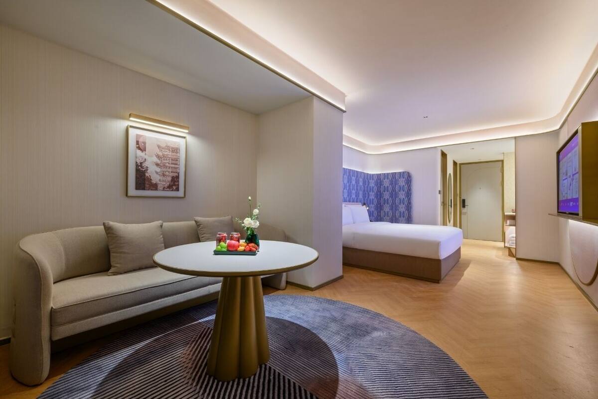 Ett modernt hotellrum med en soffa, ett runt bord med frukt, en säng och en tv.
