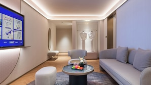 A modern hotel room with a flat-screen TV mounted on the wall, a round coffee table with fruits and a vase, a grey sofa, a grey armchair, and a small ottoman. - Manxin Hotel Wuhan Hankou Xinhua Road (Wuhan)
